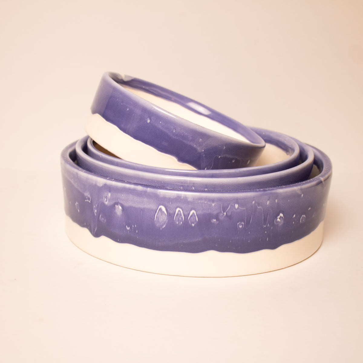 Three nested ceramic bowls with a glossy purple glazed exterior and a natural off-white clay base, photographed on a warm beige background.