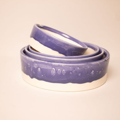 Three nested ceramic bowls with a glossy purple glazed exterior and a natural off-white clay base, photographed on a warm beige background.