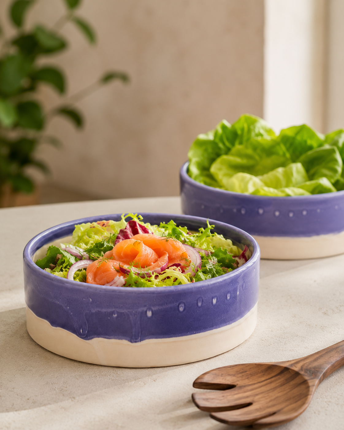 Two ceramic salad bowls with a blue glazed exterior and a beige unglazed base, shown on a tabletop. The front bowl contains a mixed salad with salmon and red onion, and the back bowl contains lettuce.