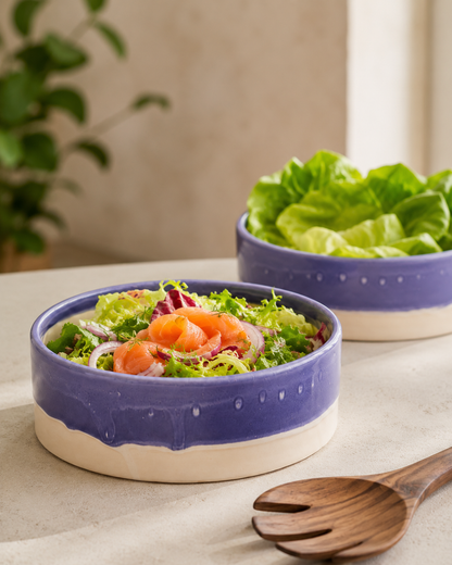 Two ceramic salad bowls with a blue glazed exterior and a beige unglazed base, shown on a tabletop. The front bowl contains a mixed salad with salmon and red onion, and the back bowl contains lettuce.