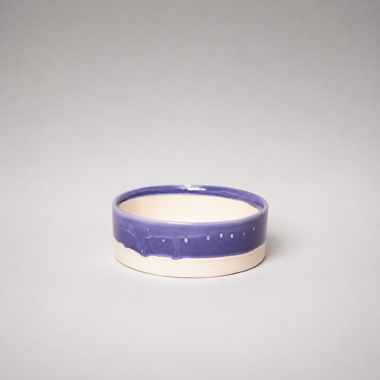 A small round ceramic bowl with a glossy purple glazed upper band and a matte off-white lower body, photographed against a neutral gray studio background.