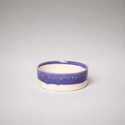 A small round ceramic bowl with a glossy purple glazed upper band and a matte off-white lower body, photographed against a neutral gray studio background.