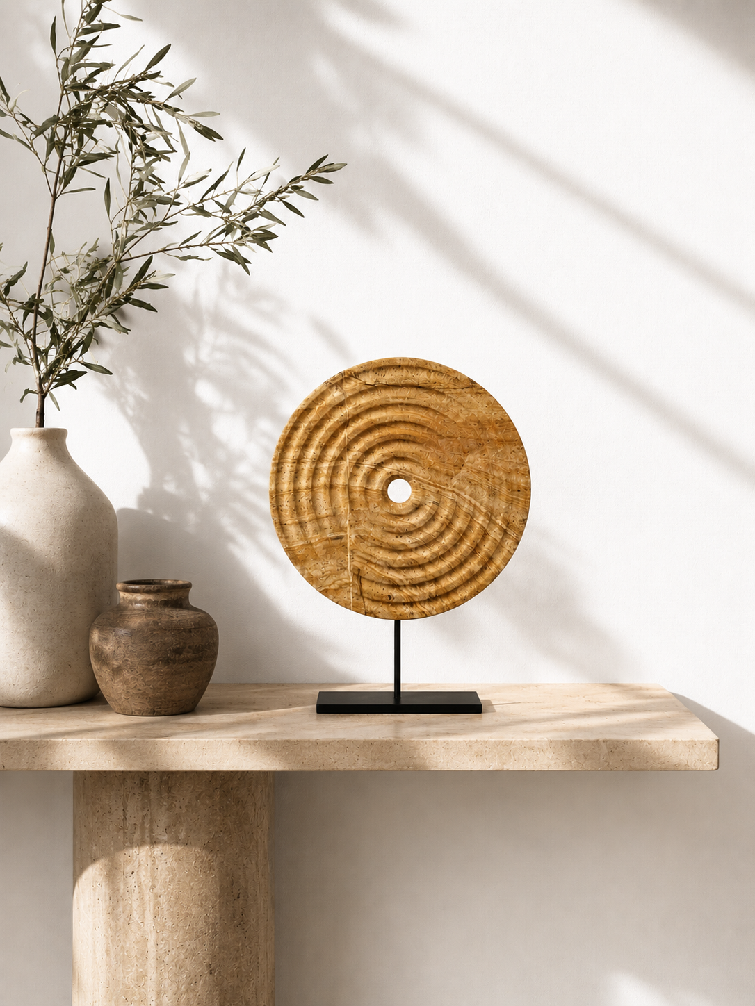 Minimal warm-toned living room interior featuring a handcrafted circular stone wall art mounted on a beige wall. Soft natural daylight enters through large windows, illuminating neutral furniture, dark wooden accents, a sculptural chair, textured curved sofa, ceramic vase with branches, and a calm modern organic aesthetic.