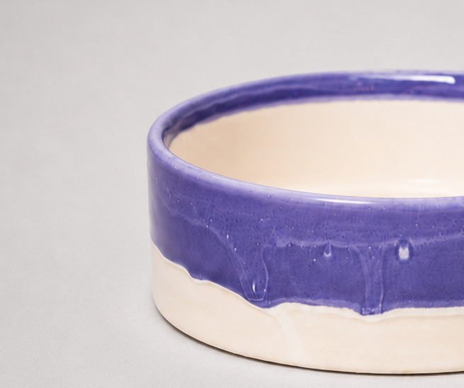 Close-up of a ceramic bowl with a glossy purple glazed rim and a matte cream-colored interior and base, set against a neutral light gray background.