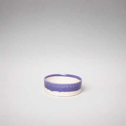 A small round ceramic bowl with a glossy purple glazed rim and a matte off-white interior, centered against a neutral gray gradient background.